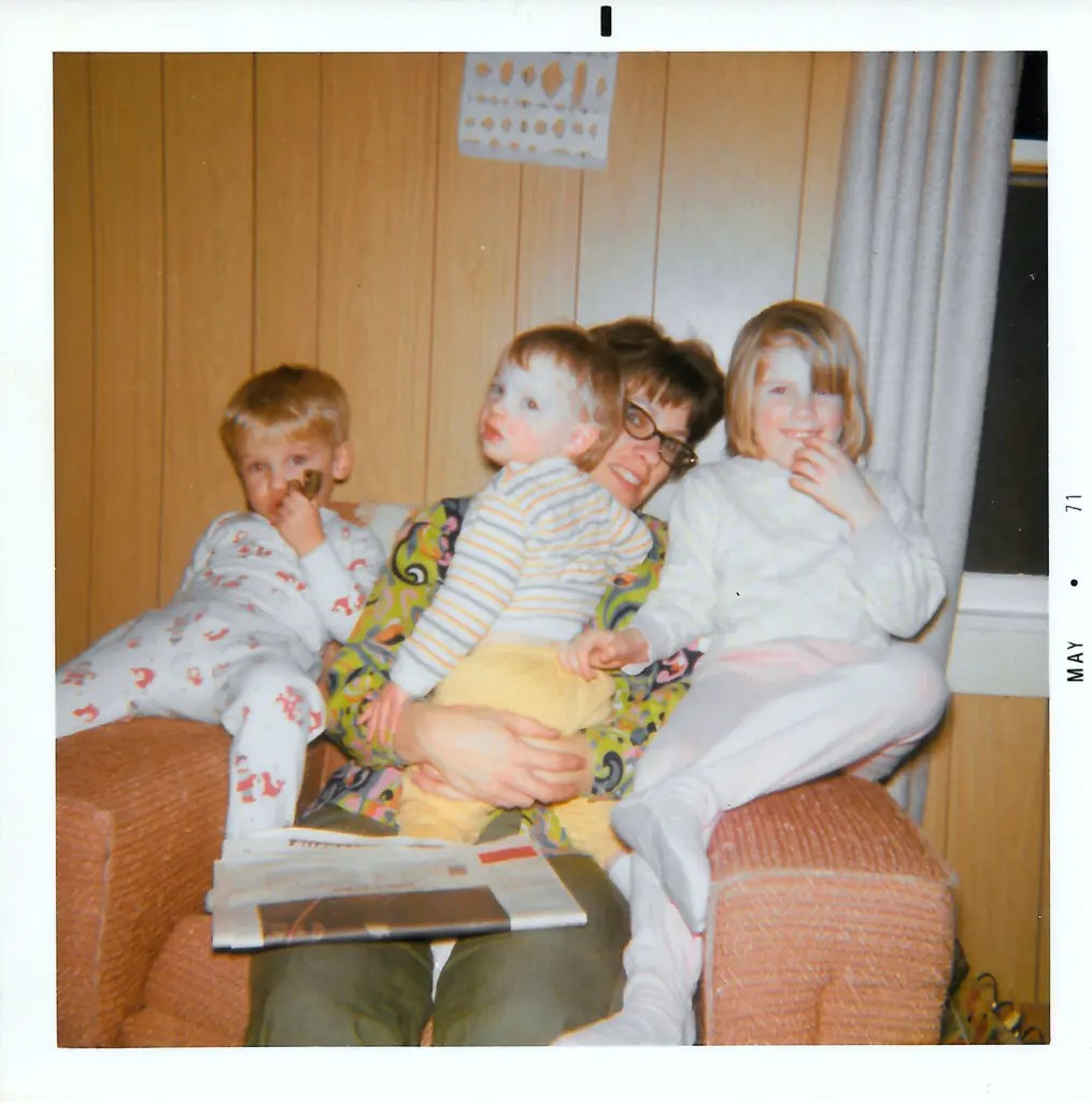 A person wearing glasses and three children sit close together in an armchair with a newspaper on the person's lap in a wood-paneled room.