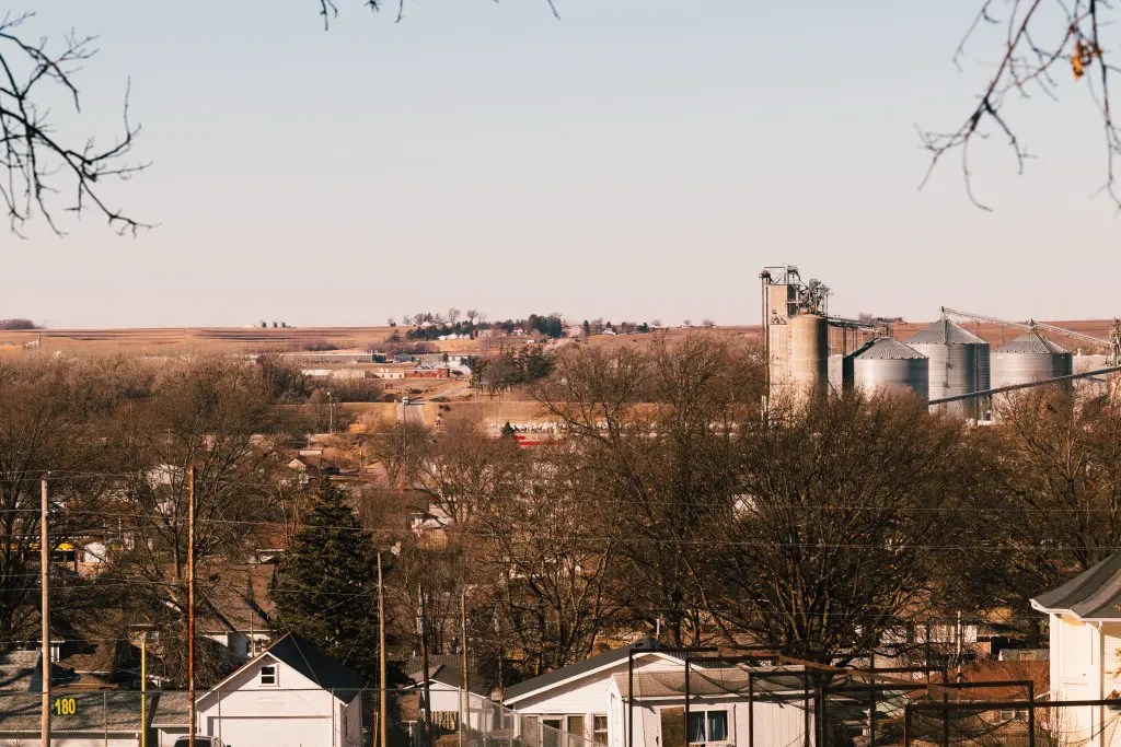 View of a small town with houses and leafless trees in the foreground and large grain silos and farm fields in the distance.