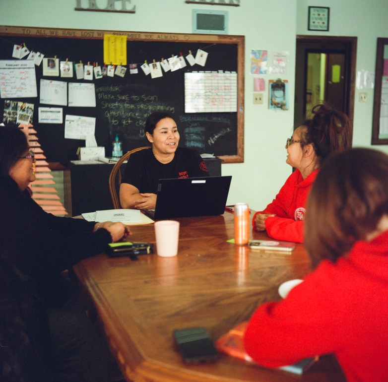 Four people sit around a wooden table with papers, drinks and a laptop in a room with a chalkboard covered in notes and photos.
