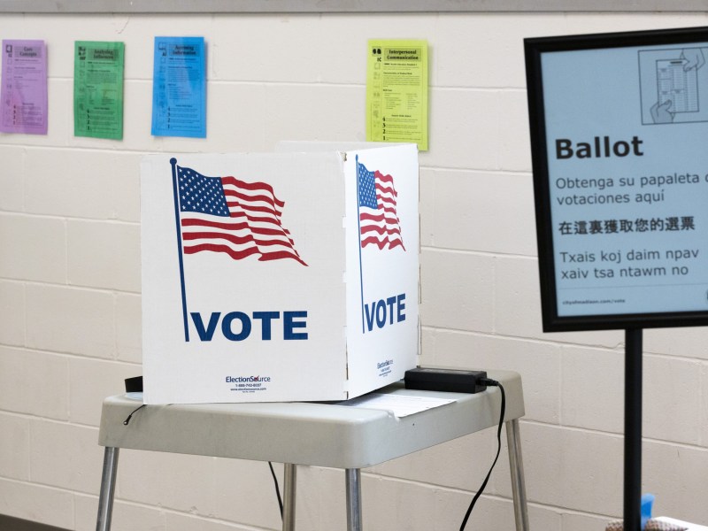 A voting station with American flag graphics and the word "VOTE" is next to a sign reading "Ballot" with instructions in multiple languages.