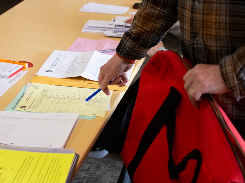 A person holding a red bag points with a pen at a piece of paper on a table covered with papers of varying colors and envelopes.