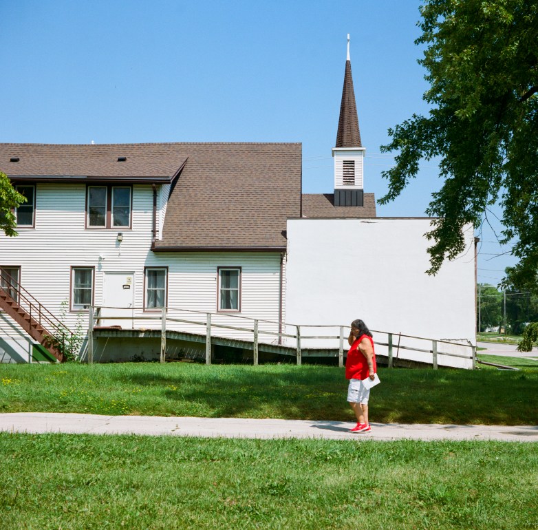 A person wearing a red shirt and white shorts walks on a sidewalk in front of a white building with a steeple and a wooden ramp.