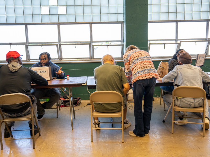 People sit across from others at tables in a room with a green wall and large windows, with signs on laptops reading "FEMA"