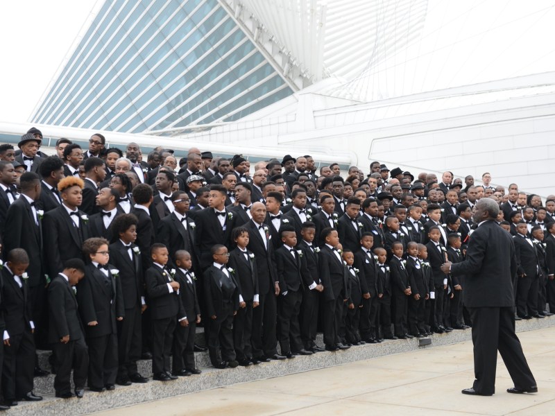 A large group of people in black tuxedos stand in rows on steps outside a modern building, while one person in a tuxedo stands in front facing them with hands raised.