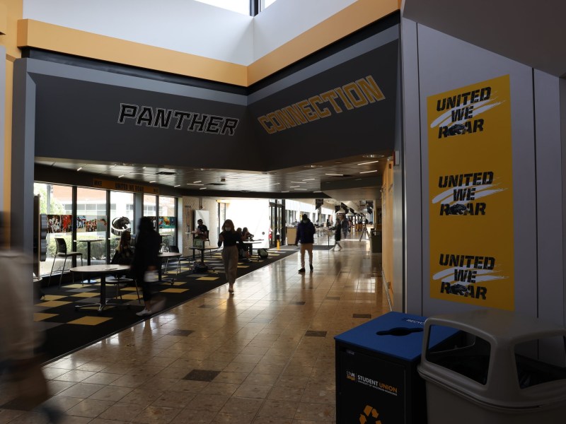 People walk in an indoor hallway with a tiled floor and signs reading "PANTHER CONNECTION" and "UNITED WE ROAR," near tables, chairs and recycling bins.