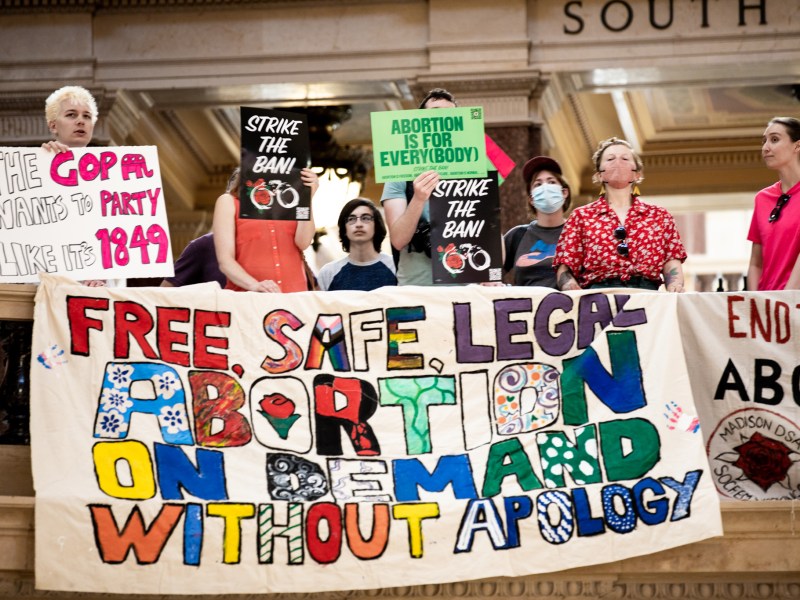 People on a marble balcony hold signs that say “FREE, SAFE, LEGAL ABORTION ON DEMAND WITHOUT APOLOGY,” “ABORTION IS FOR EVERYBODY,” “STRIKE THE BAN!” and more.