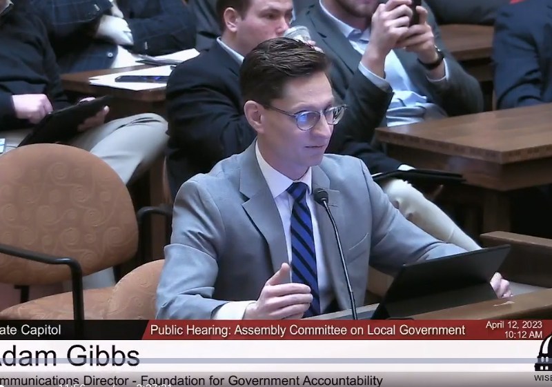 A person speaks into a microphone at a table, with a tablet in front of the person and others seated behind, as on-screen text reads “Adam Gibbs” and “Public Hearing: Assembly Committee on Local Government”
