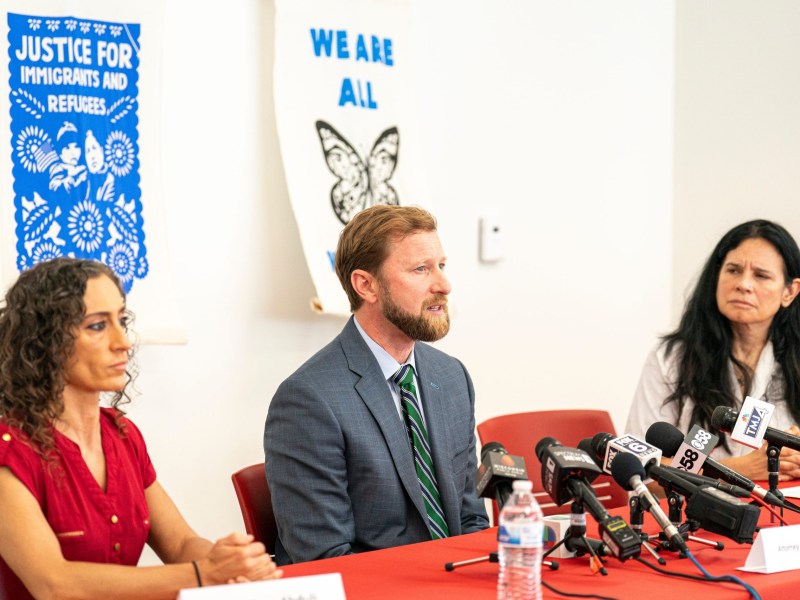 Three people are seated at a table with microphones perched atop.