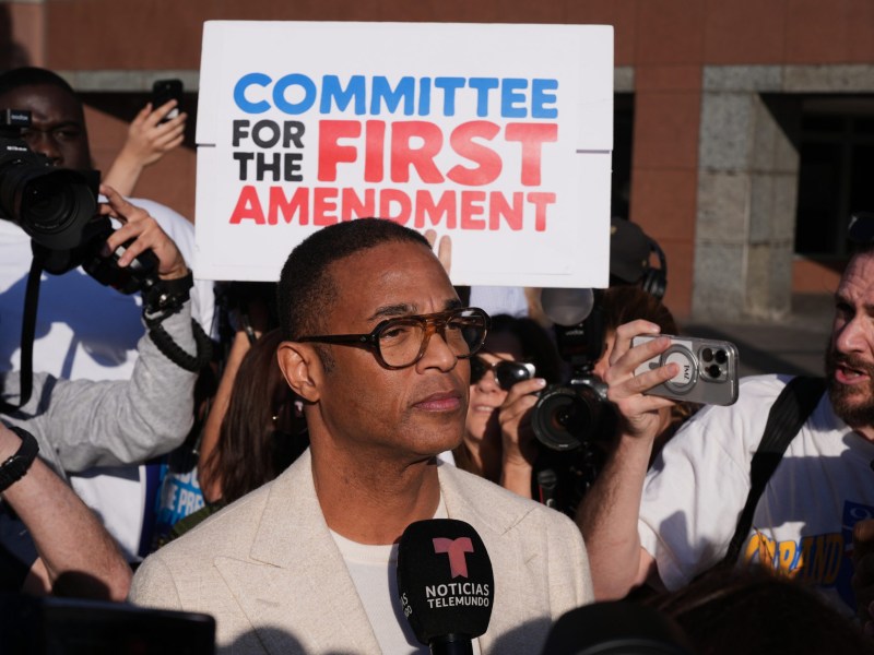 A man stands in front of a microphone with people in the background and a sign reading "Committee for the First Amendment."