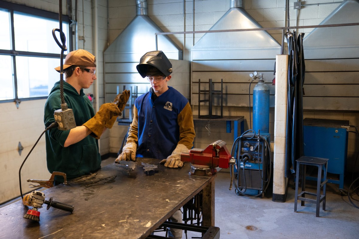 Two people wearing safety glasses and gloves stand at a metal worktable with cut metal pieces and tools in a room with a garage door to the left. Other equipment is in the background next to a wall.