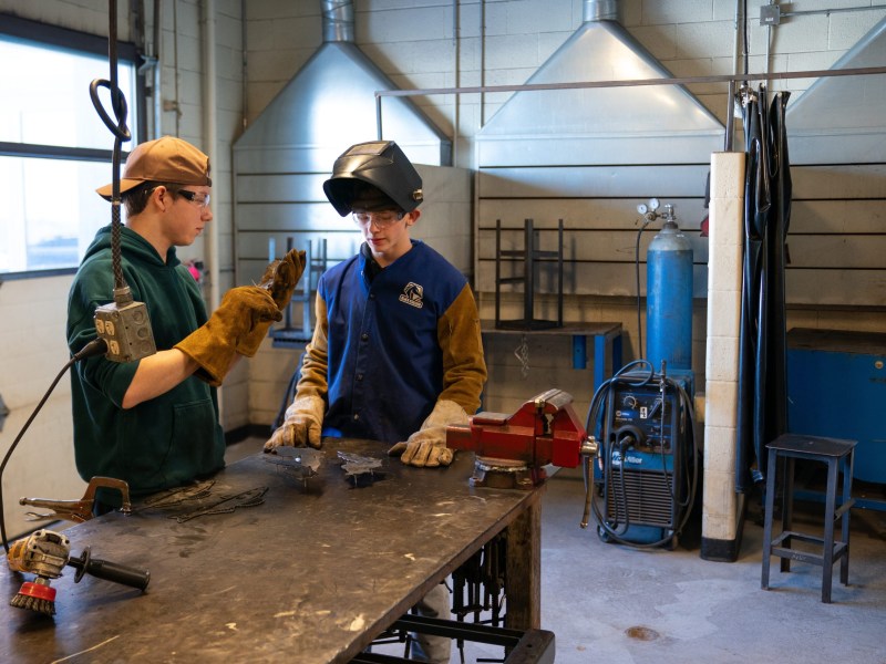 Two people wearing safety glasses and gloves stand at a metal worktable with cut metal pieces and tools in a room with a garage door to the left. Other equipment is in the background next to a wall.