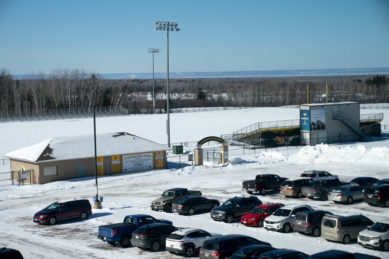 A snow-covered football field and bleachers are behind a parking lot filled with cars. A building next to the football field entrance has a sign that says "Northwestern Tigers State Champions 1988"