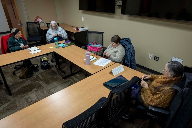 People sit around tables knitting with needles and yarn inside a room, with papers, bags, water bottles, and other items on the tables.