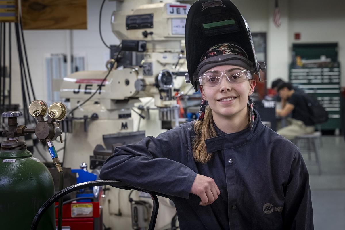 A person wearing safety glasses and a raised welding helmet leans an arm on a machine in a workshop, with industrial equipment and another person working in the background.