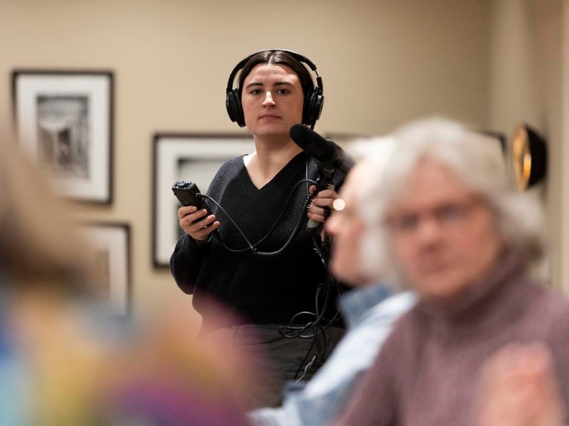 A reporter wearing headphones holds a microphone and recording device while standing in a room with framed pictures and people in the foreground.
