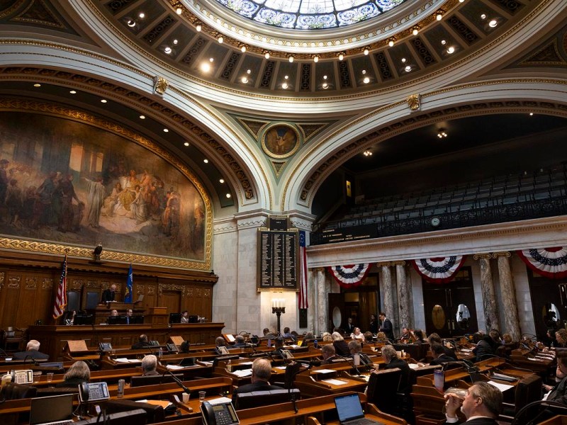 People sit at desks inside an ornate room beneath a domed ceiling, with U.S. and state flags, a large mural and an electronic board visible above the floor.