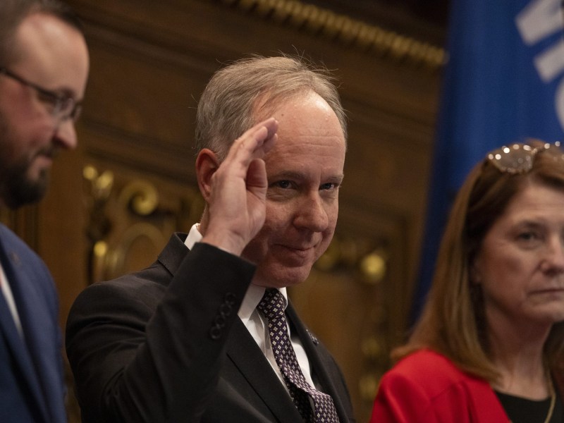 A person in a suit raises a hand to the forehead while standing between two other people in a wood-paneled room with a blue flag in the background.