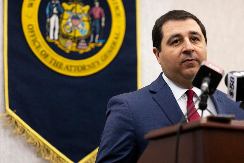 A person stands at a podium near microphones with a banner behind them displaying the Wisconsin state seal and the words "Office of the Attorney General."