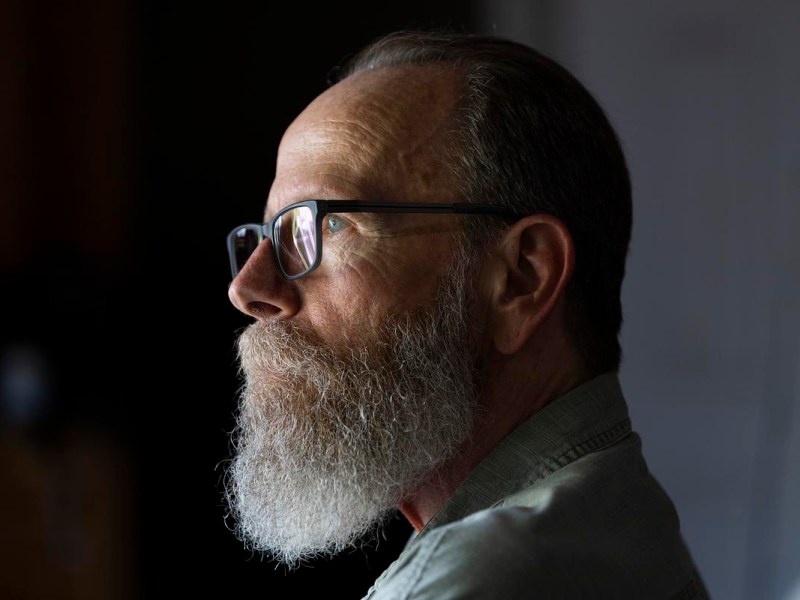 A person with glasses and a long beard faces sideways in soft light against a dark background.