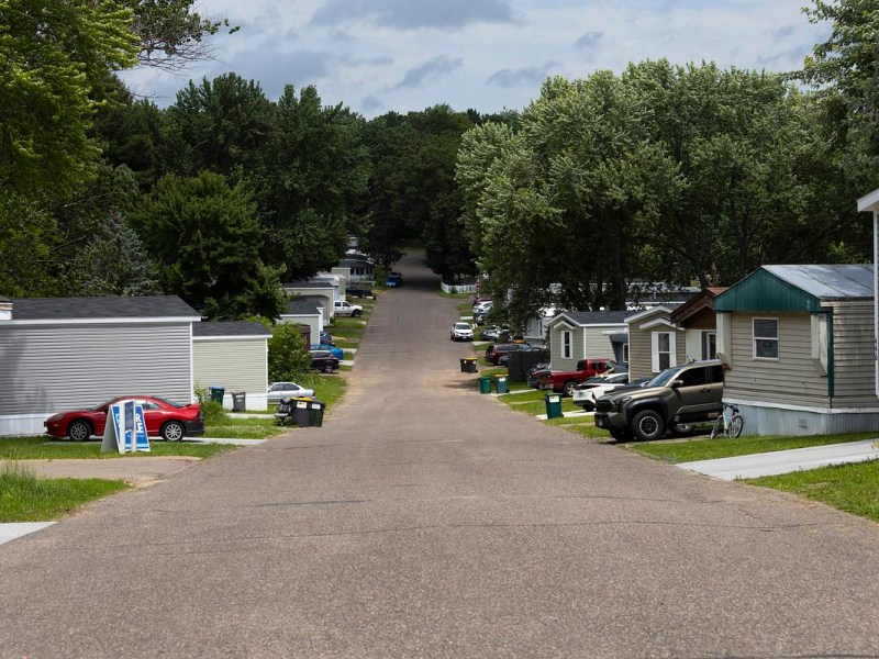 Paved road lined with manufactured homes, parked cars and trees under a cloudy sky
