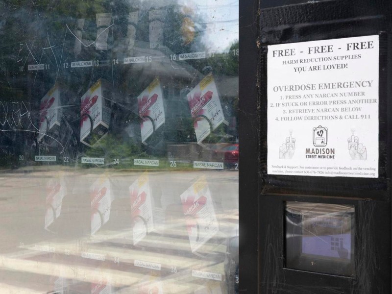 A man with light brown hair pulled back stands outside on a sunny day, leaning against a railing. He wears a light blue button-up shirt with a subtle dot pattern and has a small earring in his left ear. In the background are trees, an apartment building, and a tan residential structure with a gray roof.