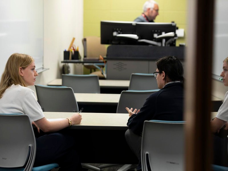 Two people sit across from each other at one of three tables in a room while a person stands next to a computer monitor in the background.