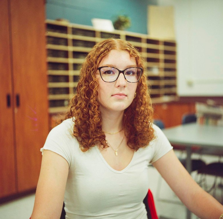 A person wearing glasses and a white shirt sits in a room with tables and mail slots in the background.
