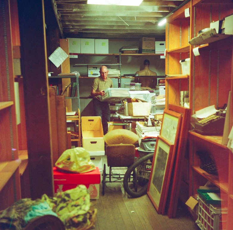 A person stands in a cluttered room filled with boxes, papers and framed items, holding a stack of items.
