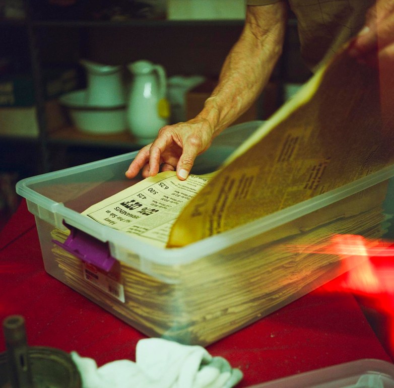 A person’s hands sort through old newspapers stored in a clear plastic bin on a table.