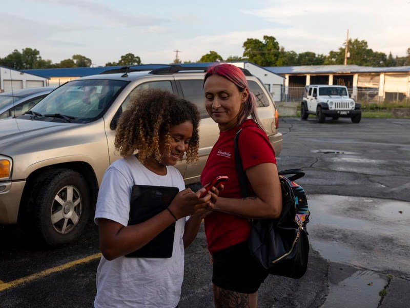 Two people stand in a parking lot as one looks at a phone and holds a tablet while the other stands nearby with a backpack.