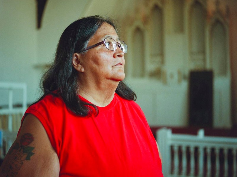 A person wearing glasses and a red sleeveless shirt stands near white railings with out-of-focus arched architectural details in the background.
