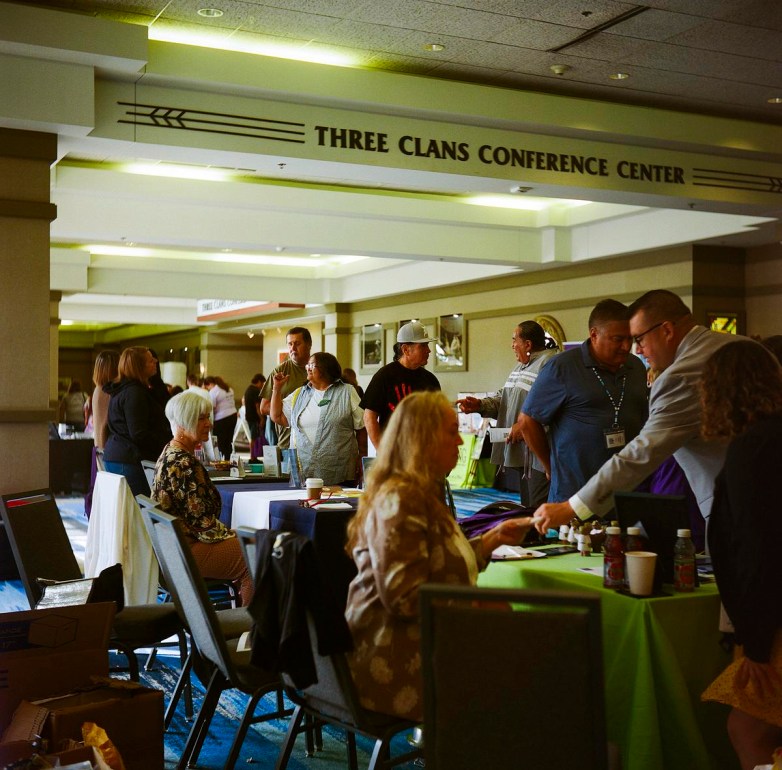 People stand and sit at tables in a hallway under a sign reading "Three Clans Conference Center"