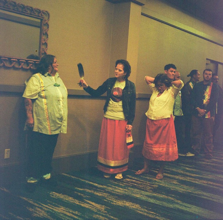 People stand in a hallway, including one person holding a feather toward another wearing a shirt with a green turtle decoration, while others wait nearby.