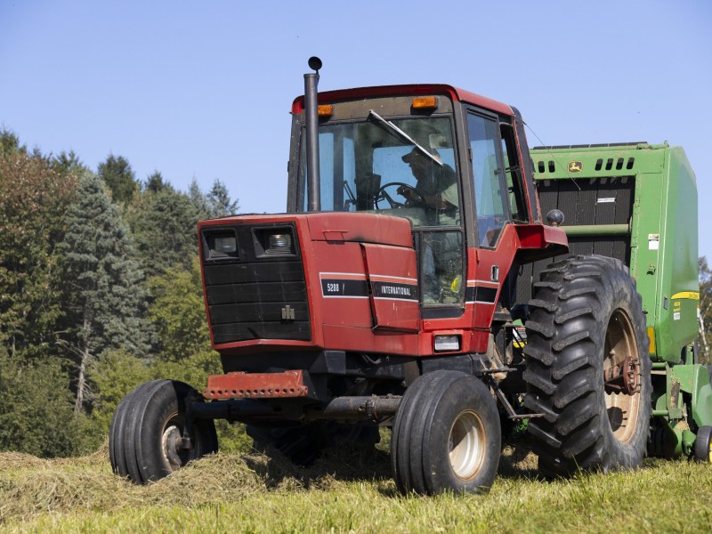 A red International tractor pulls green farm equipment across a field, with trees in the background and a person visible holding a steering wheel inside the tractor.