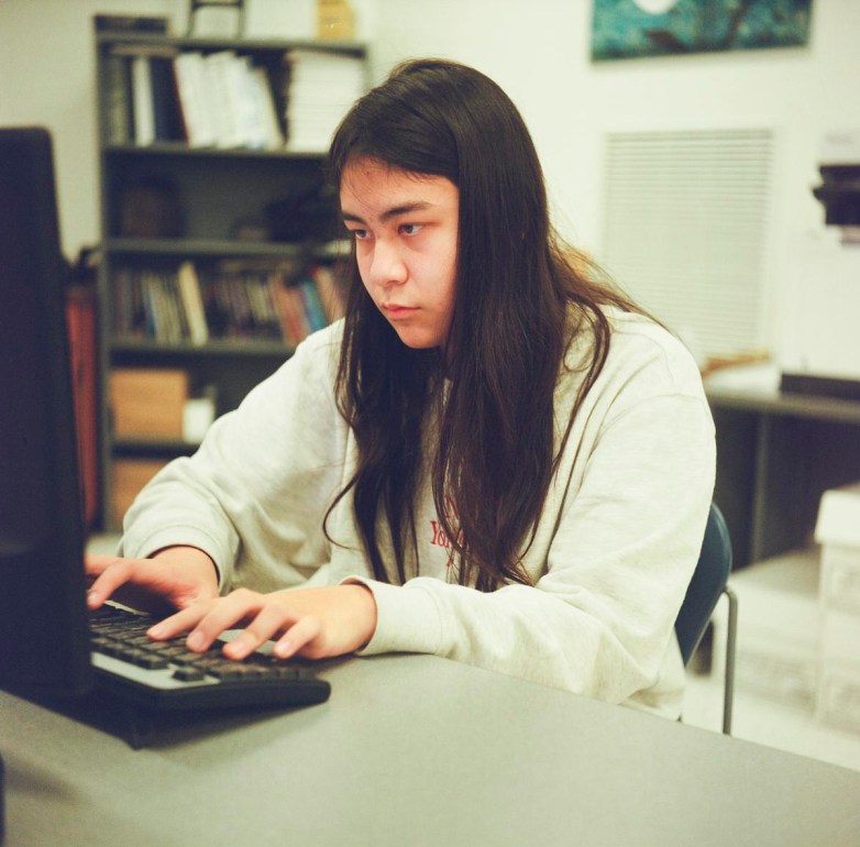 A person wearing a light sweatshirt types on a computer keyboard in a room with shelves and books in the background.