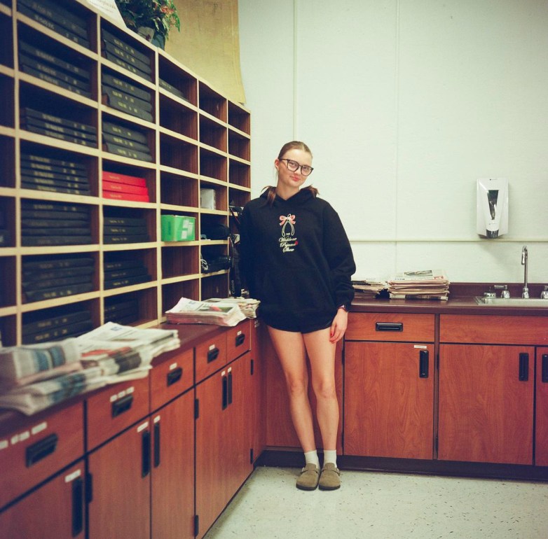 A person wearing a black hoodie and glasses stands beside shelves filled with bound volumes. Stacks of newspapers are on a counter.