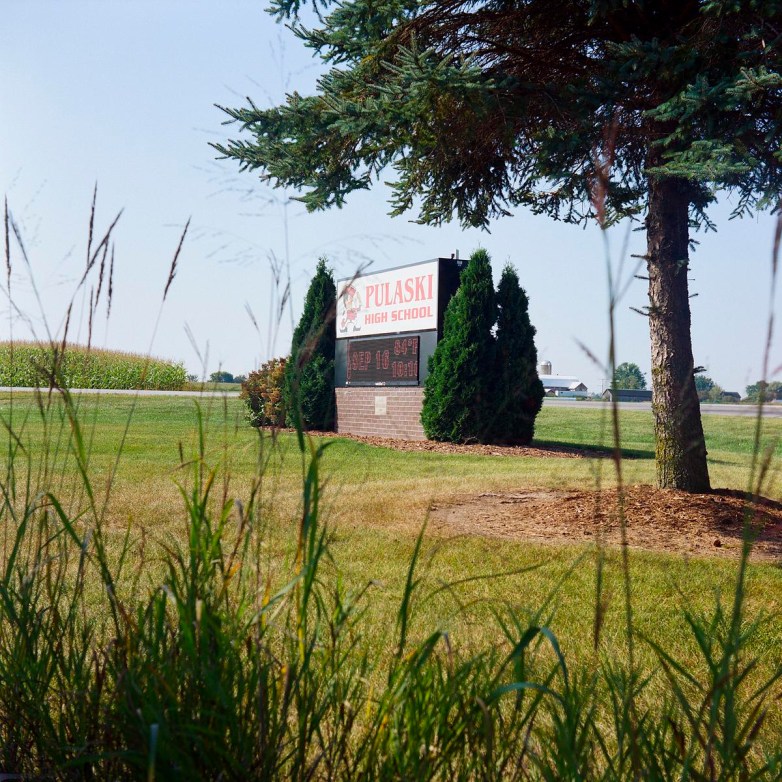 A sign reading "Pulaski High School" stands beside grass and a large tree, with a cornfield visible in the background.
