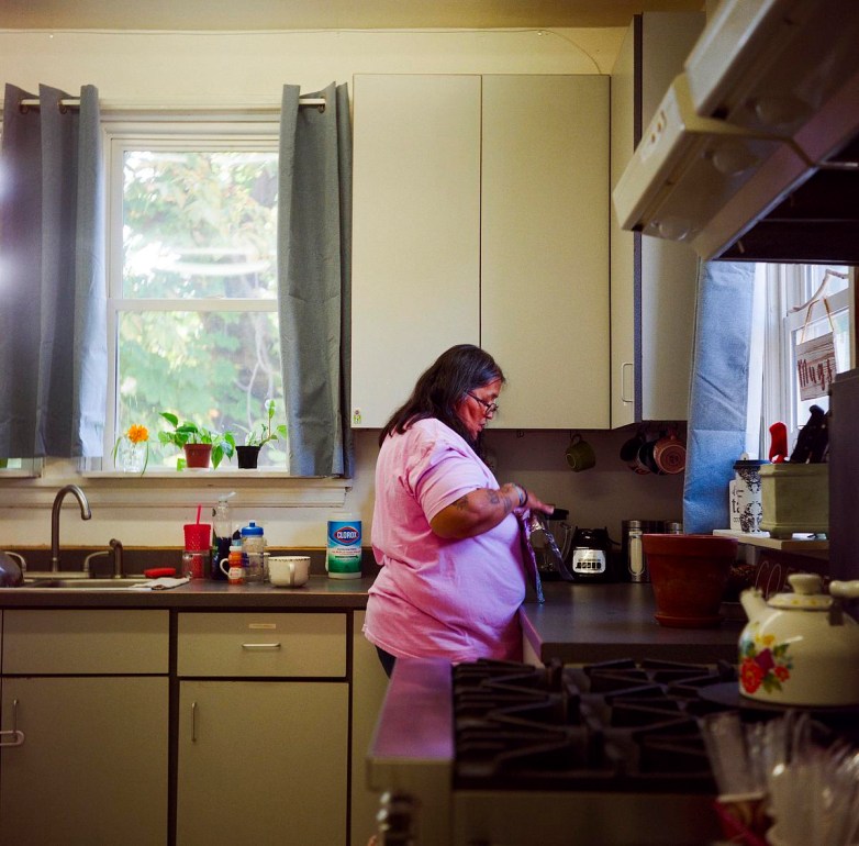 A person wearing a pink shirt stands at a kitchen counter near a window with potted plants on the sill.