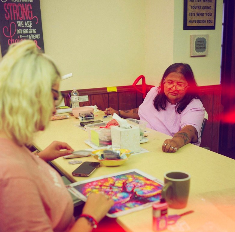 Two people sit at a table filled with colorful craft projects, supplies, mugs, and art materials.