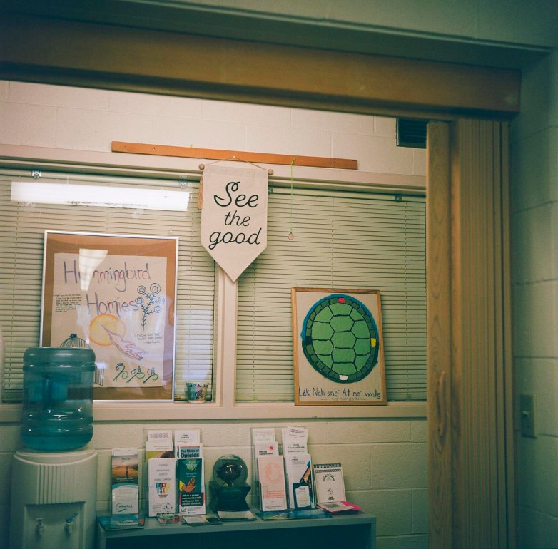 Posters and a banner reading "See the good" are on a wall with windows with shades above a water dispenser and pamphlets on a table.