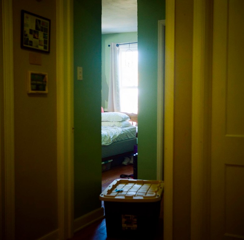 Sunlight filters through a window into a bedroom with a bed, seen from a hallway with a plastic storage bin on the floor.