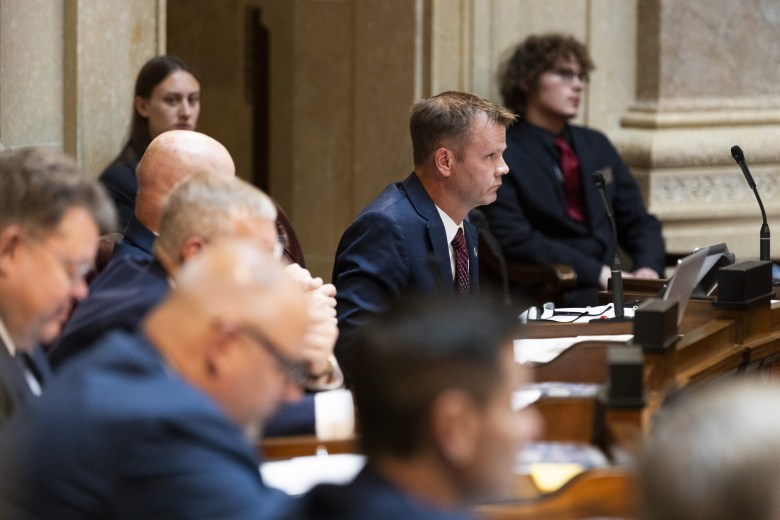 A person in a blue suit sits at a desk with a microphone and laptop in a room while others sit nearby.