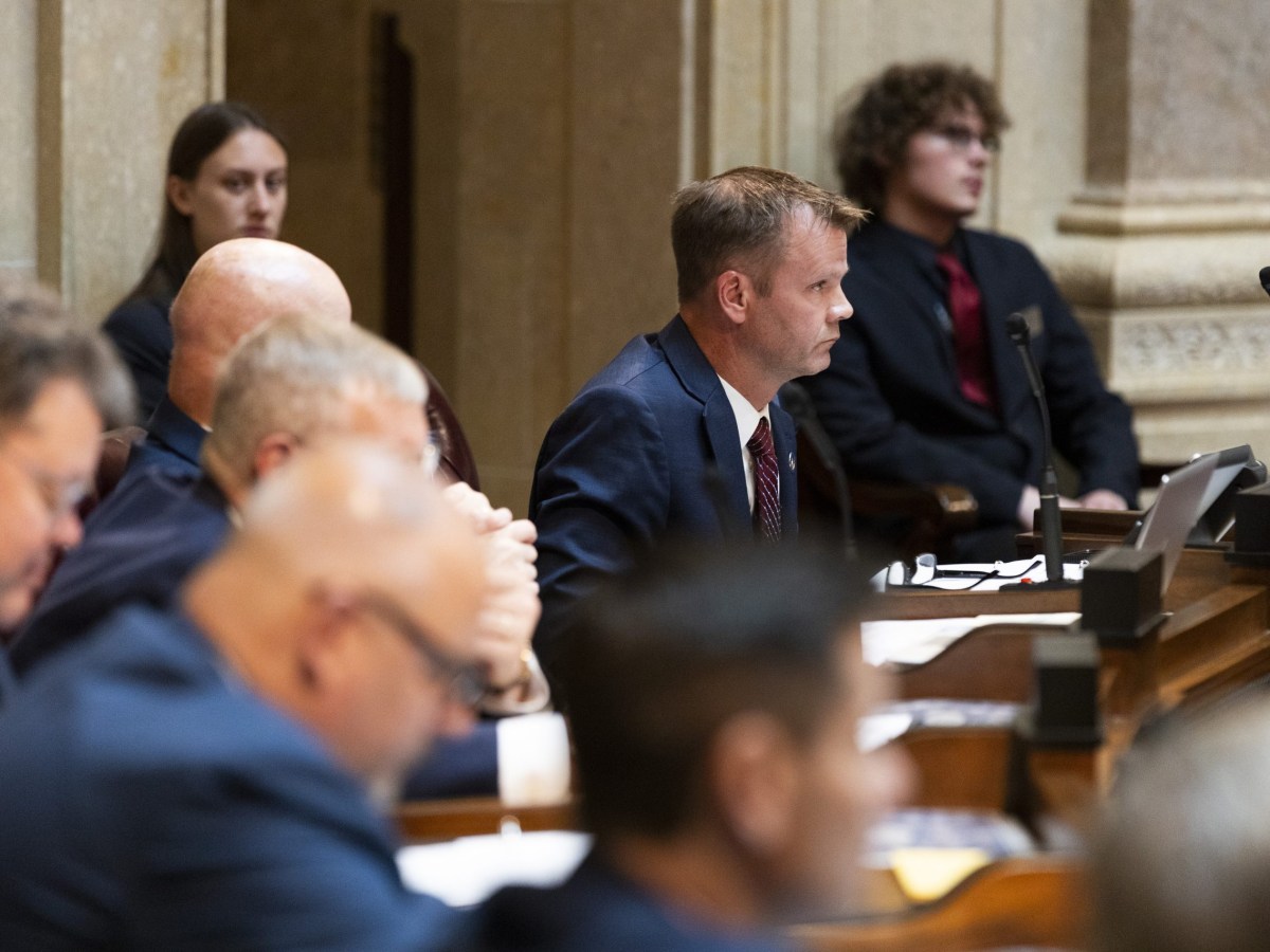 A person in a blue suit sits at a desk with a microphone and laptop in a room while others sit nearby.