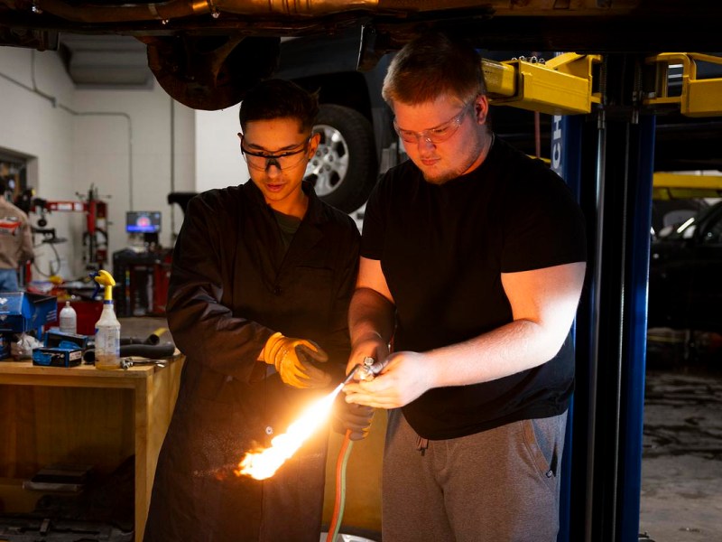 Two people wearing safety glasses stand under a vehicle lift as one holds a torch emitting a bright flame, with tools and equipment in the background.