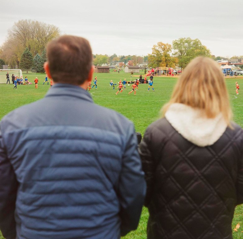 Two people are seen with their backs to the camera watching a youth soccer game on a grassy field, where players in blue and orange uniforms run with a goal to the left.