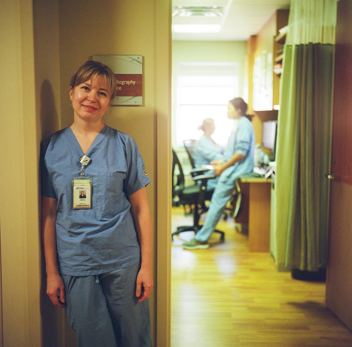 A person wearing blue scrubs and an ID badge stands beside a doorway, with two other people in scrubs seated at desks in a room behind the open door.