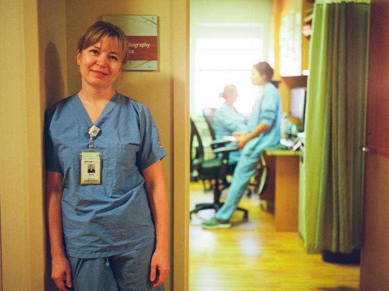 A person wearing blue scrubs and an ID badge stands beside a doorway, with two other people in scrubs seated at desks in a room behind the open door.