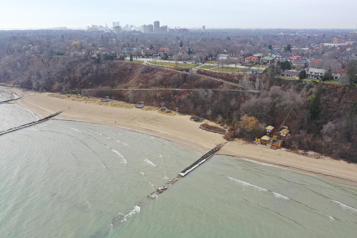 An aerial view shows a sandy beach and greenish lake water with a wooden breakwater, a wooded bluff behind the shore, houses along the top, and a small wooden structure near the sand.