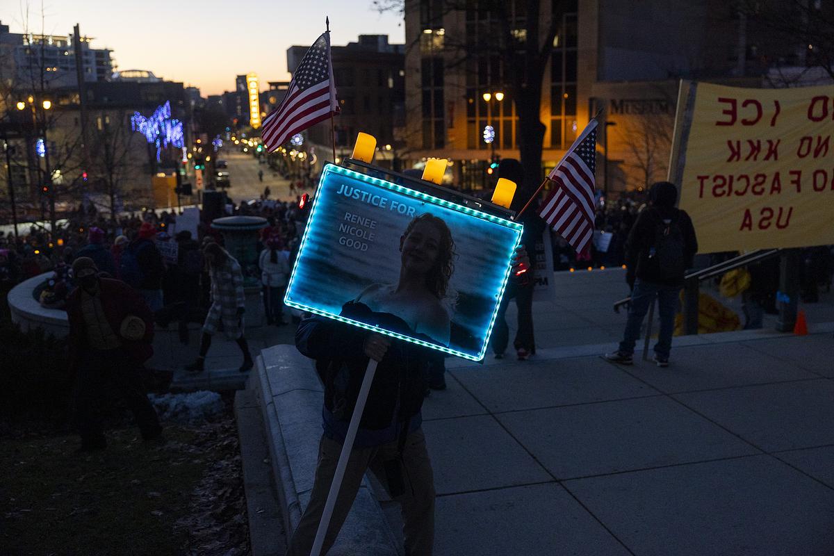 A person holds a sign that says "Justice for Renee Nicole Good" that has candles and American flags attached. Other people walk in the background.