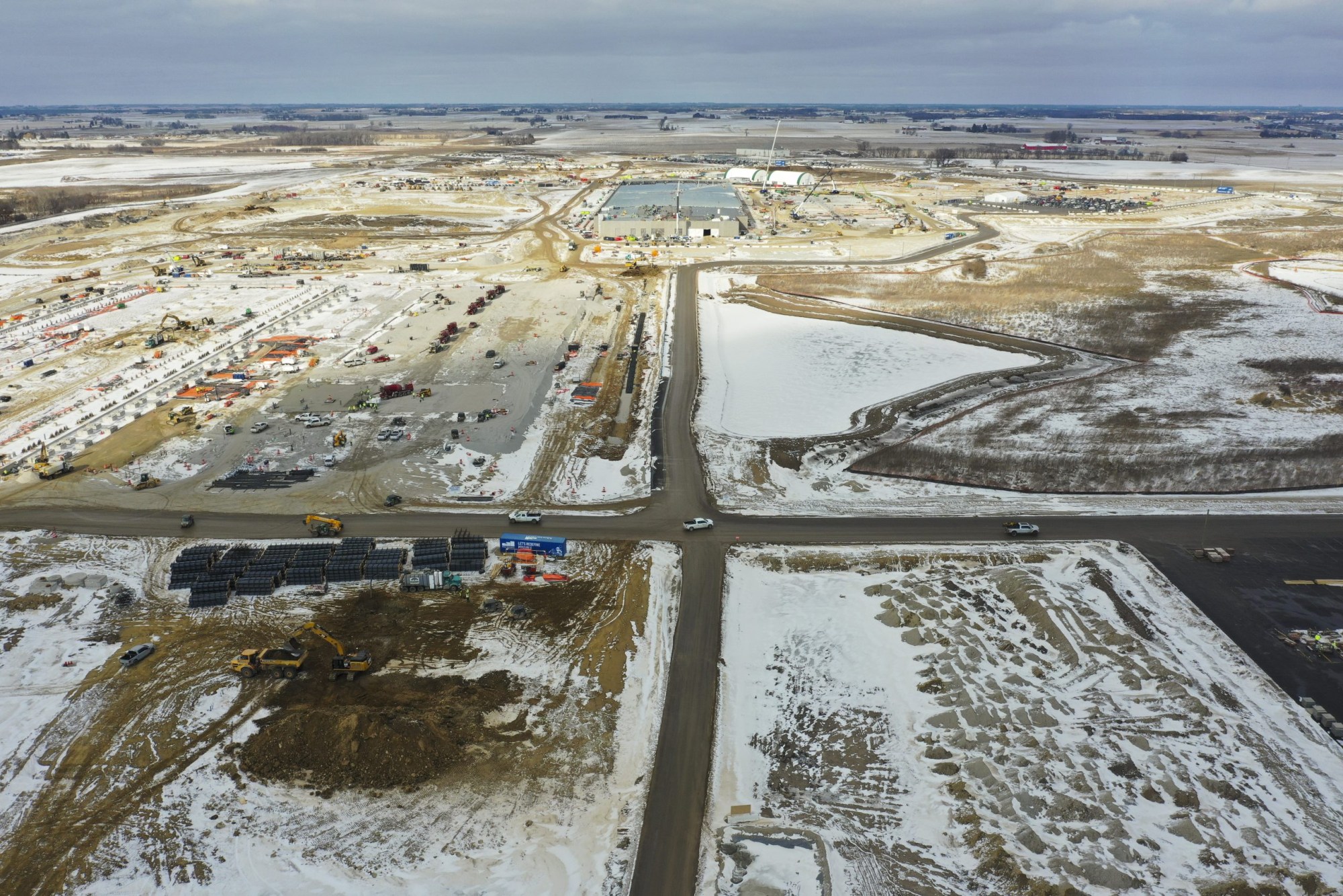 An aerial view shows a large construction site with cranes, heavy equipment and materials surrounded by snow-covered fields and intersecting roads.
